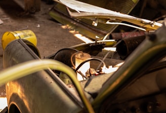 A dismantled and abandoned vehicle is situated in a dimly lit garage. The vehicle's hood is removed, exposing its internal components. Rusty barrels and a bench are in the background, along with other discarded items. Sunlight filters in, casting shadows across the dusty floor.